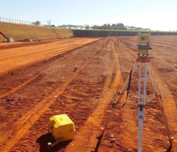 Estação total instalada em tripé realizando medições topográficas em área de terraplenagem, com solo avermelhado e superfície nivelada para preparação de obra civil.
